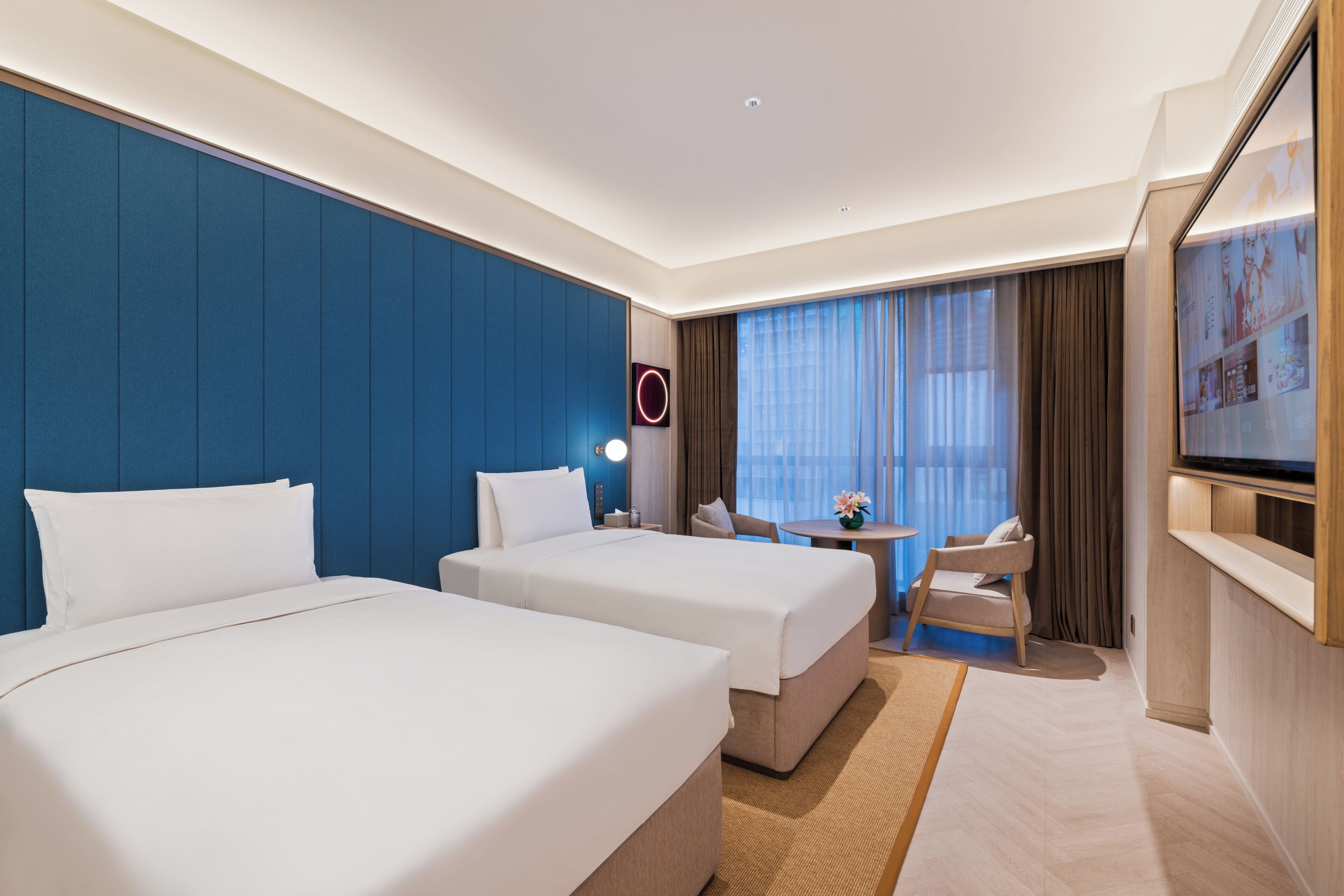 Habitació d'hotel amb dos llits, un escriptori, una cadira, una televisió i una finestra gran amb cortines.
