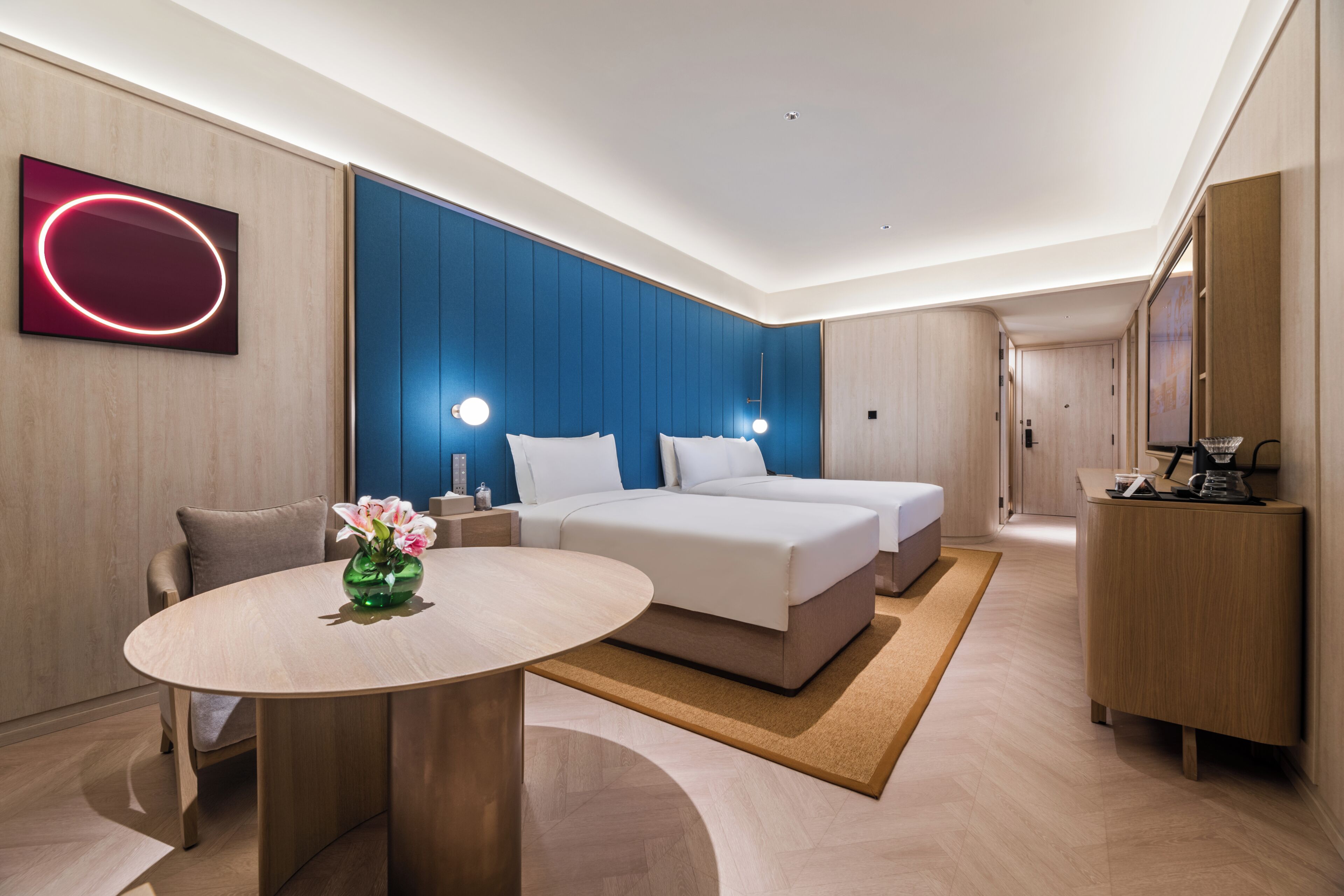 A modern hotel room with a bed, bedside tables, a round table with a vase, and a large mirror.