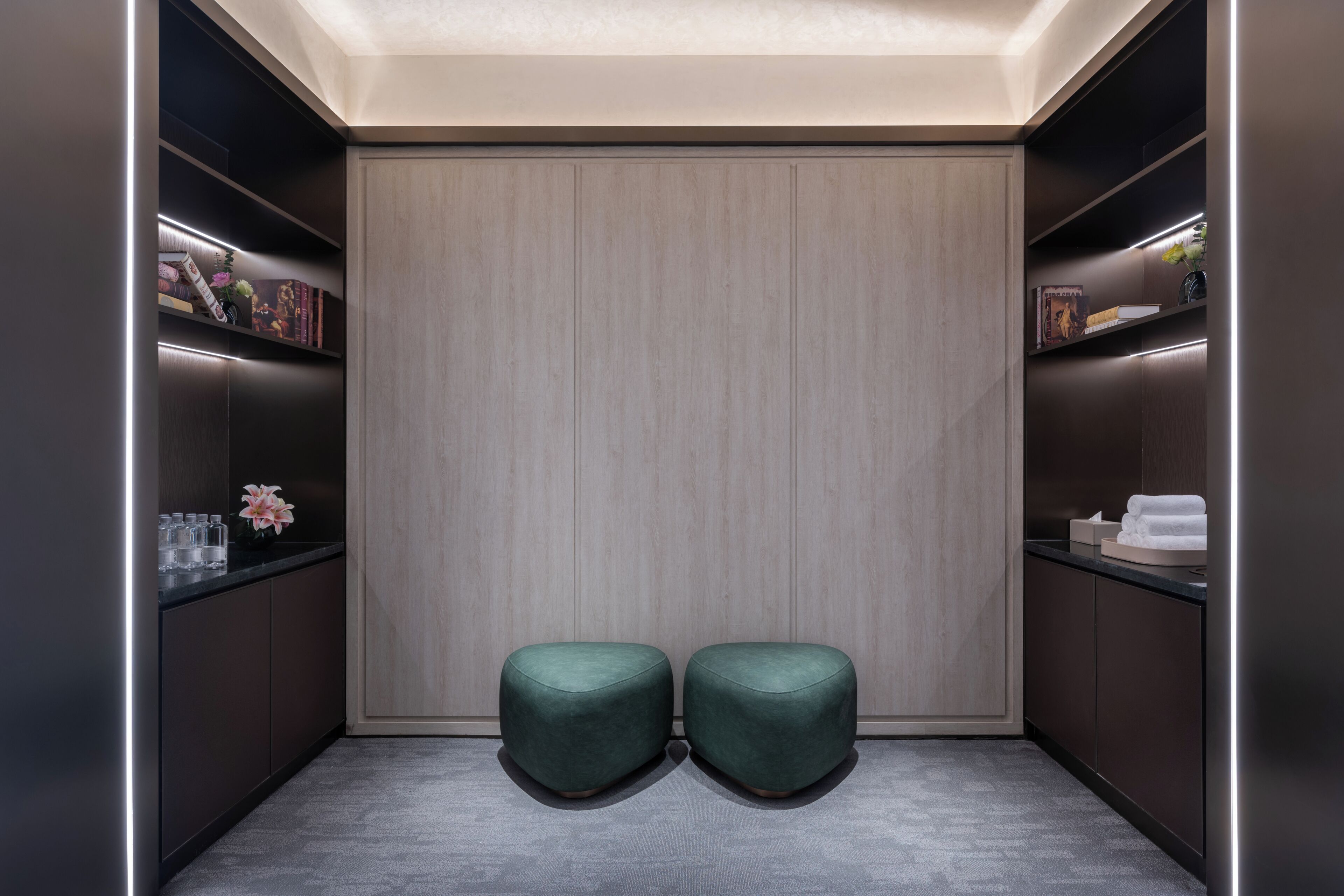Um quarto moderno, com iluminação suave e cores neutras, destacando um grande painel de madeira, duas áreas de estar com assentos acolchoados em verde e prateleiras embutidas com diversos objetos.