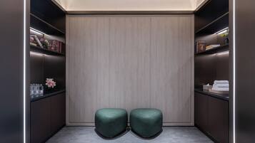 Um quarto moderno, com iluminação suave e cores neutras, destacando um grande painel de madeira, duas áreas de estar com assentos acolchoados em verde e prateleiras embutidas com diversos objetos.