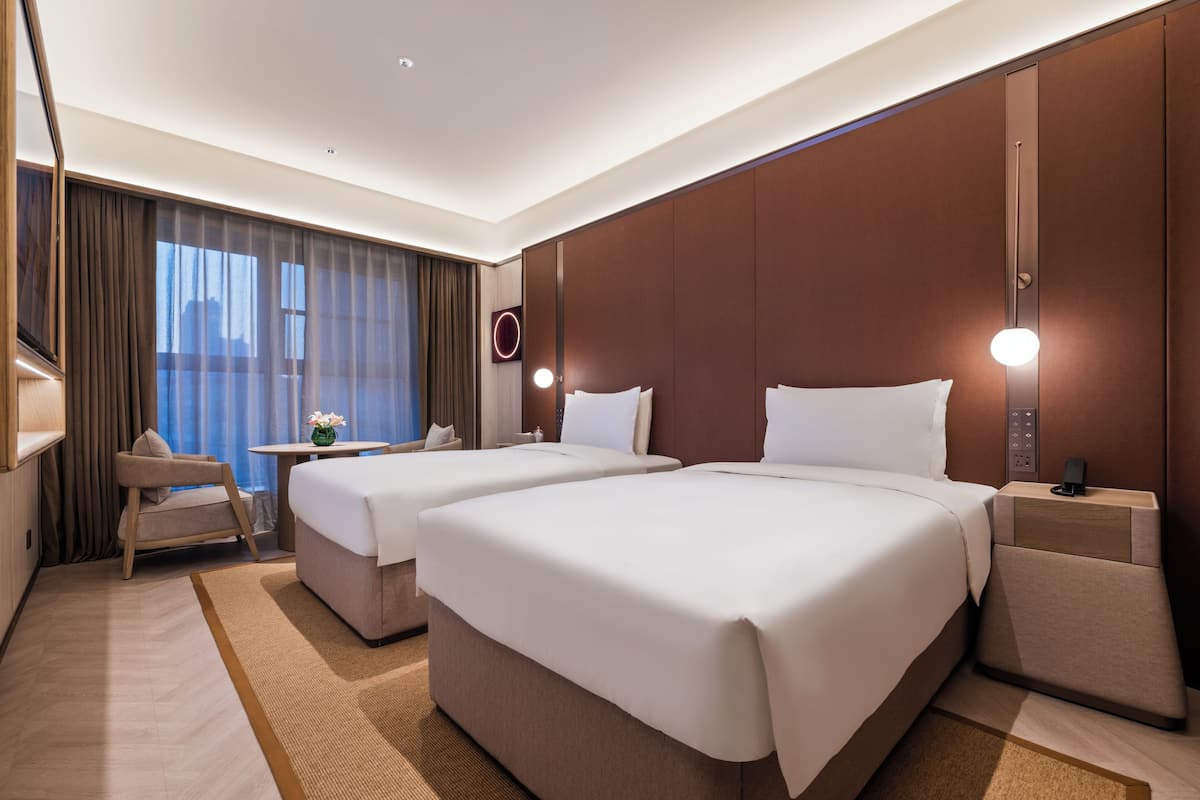 Habitación de hotel con dos camas, un escritorio, una silla, una mesita con un florero y vistas a la ciudad.