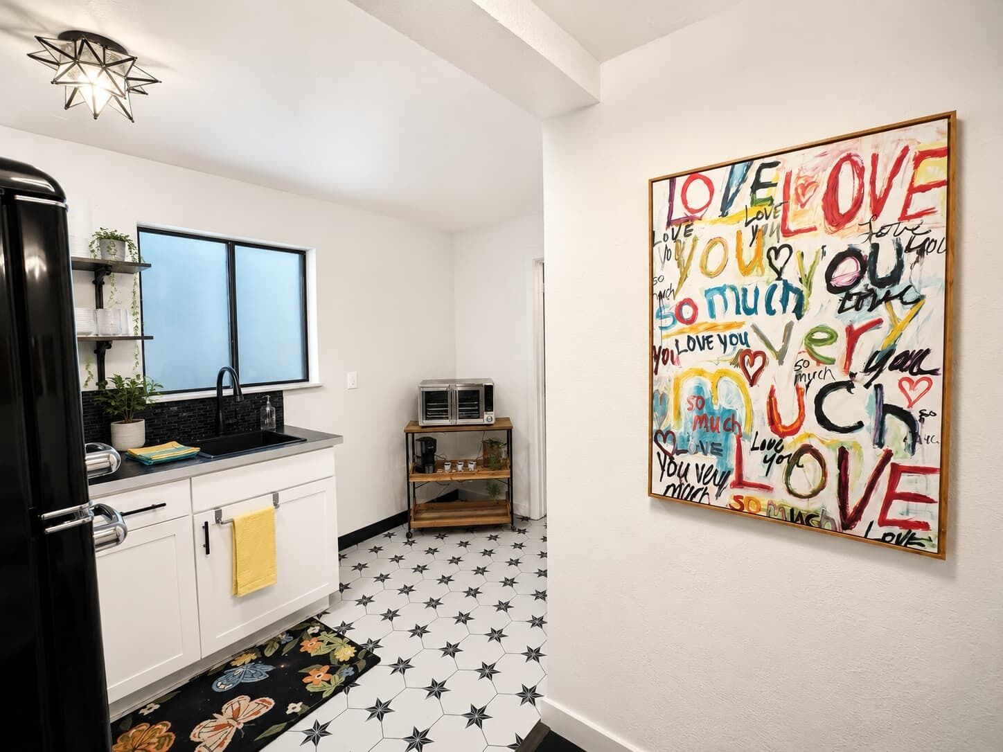 Een keuken met witte kastjes, een zwarte koelkast, een dubbele spoelbak en een magnetron. De vloer heeft een stermotief en op de muur hangt een kleurrijk kunstwerk met de tekst 'LOVE YOU'.