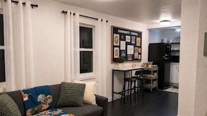 Ein Wohnzimmer mit einer Couch, einem Couchtisch und einem Essbereich mit Stühlen. Im Hintergrund ist eine Küche zu sehen.
