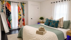 Ein Schlafzimmer mit Bett, Kissen, einem Nachttisch, einer Pflanze und Vorhängen.