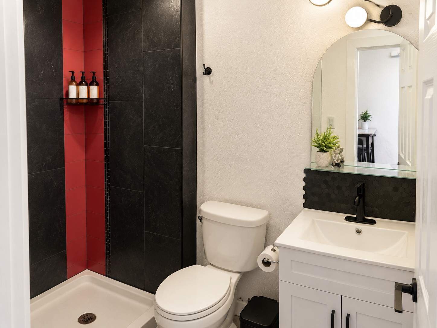 Um banheiro com vaso sanitário branco, box de chuveiro em vidro, parede com azulejos pretos e vermelhos, pia branca com torneira preta e um espelho acima da pia.