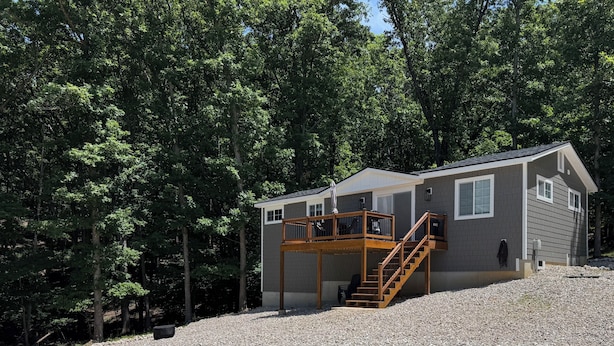 A two-story cabin with a wooden deck and stairs, surrounded by dense forest.
