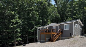 A two-story cabin with a wooden deck and stairs, surrounded by dense forest.