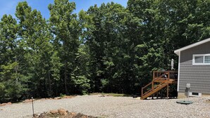 A house with a wooden staircase leading to a deck, surrounded by a gravel driveway and dense forest.