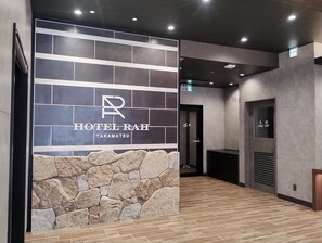 A modern hotel lobby with a stone accent wall, a sign for HOTEL RAH TAKAMATSU, and a wooden floor.