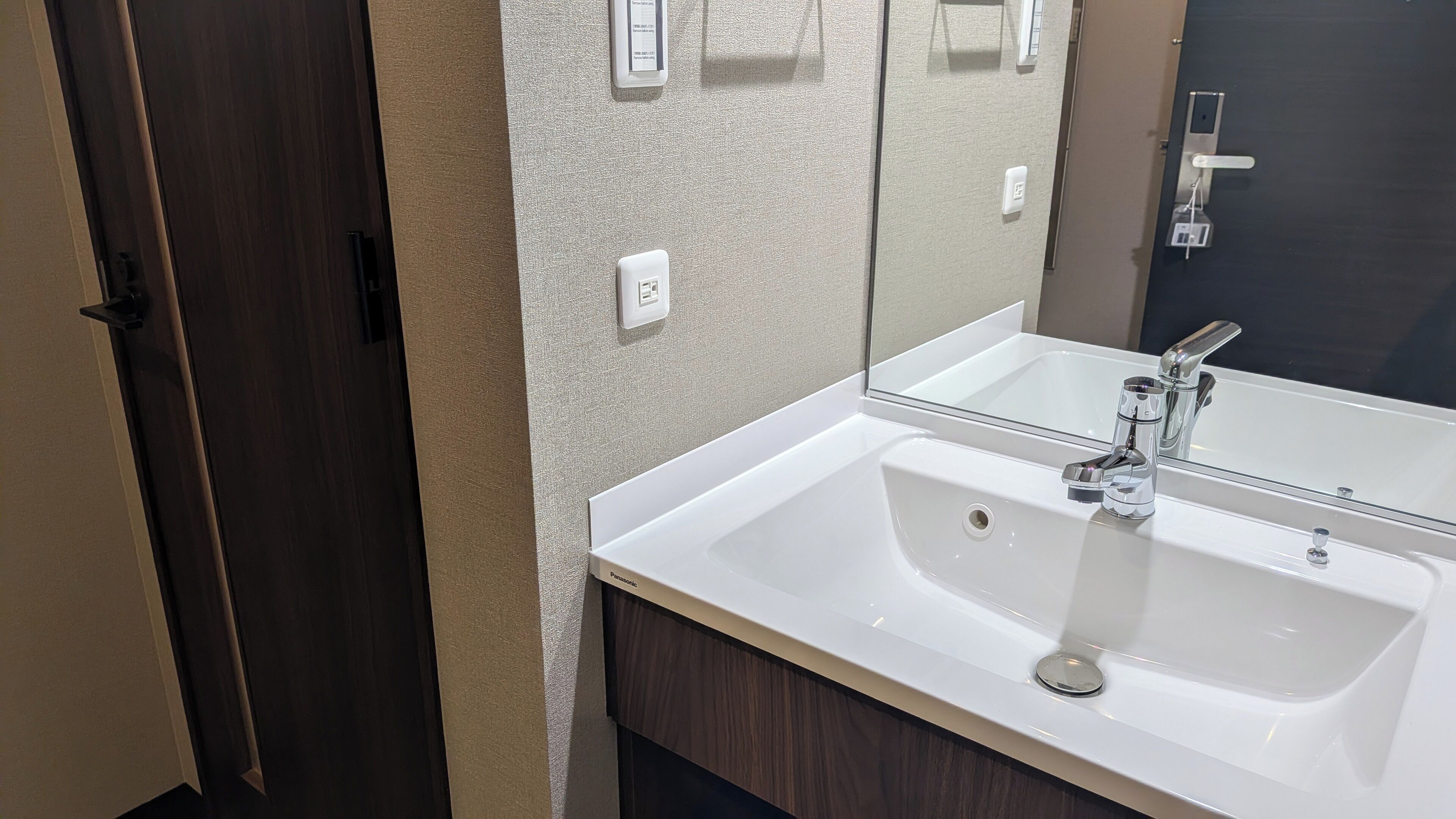 Un baño con un lavamanos blanco, un espejo, una puerta de madera oscura y una pared con textura.