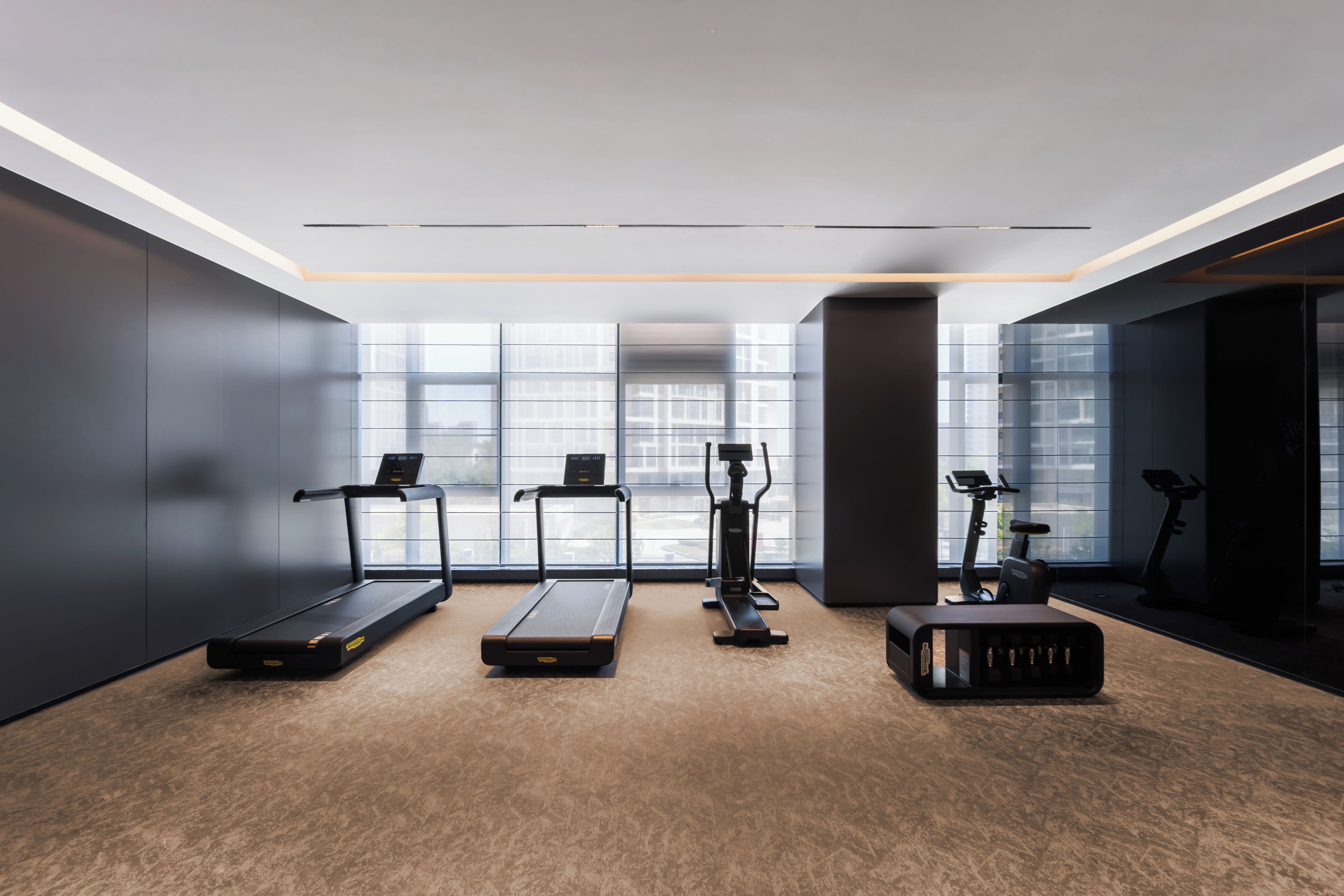 A modern gym with treadmills, an elliptical, and a stationary bike, large windows, and a minimalist design.