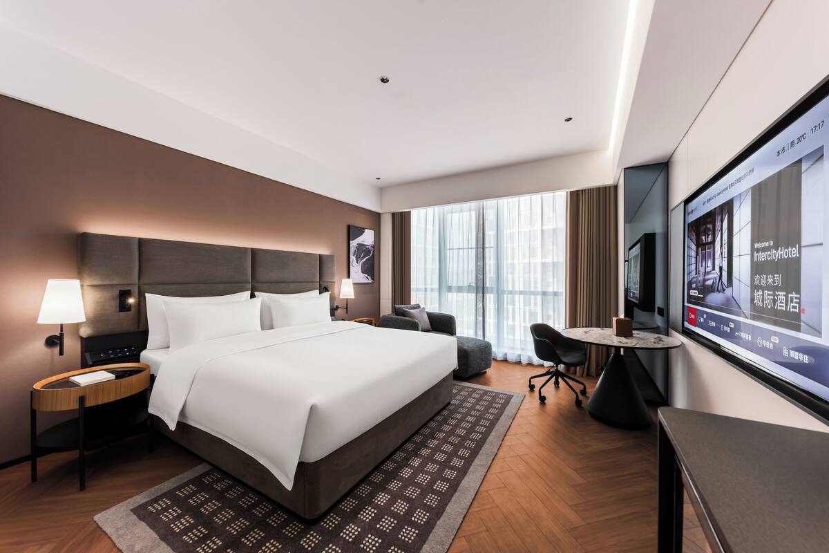 A modern hotel room with a large bed, a desk, a chair, a TV, and a window with curtains.