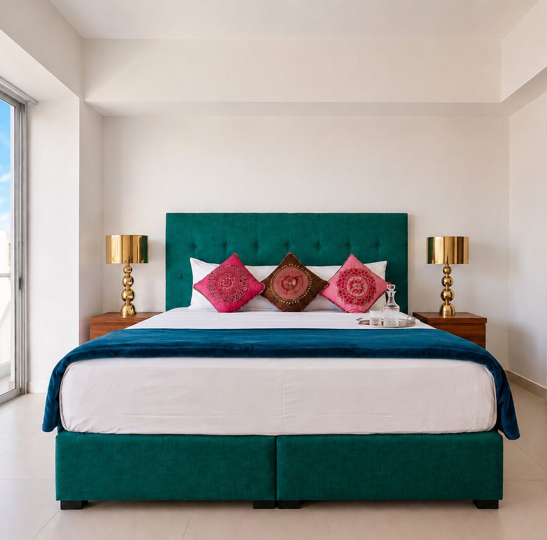 Een bed met een blauwgroen hoofdeinde, witte beddengoed en decoratieve kussens. Er staan twee nachtkastjes met lampen.