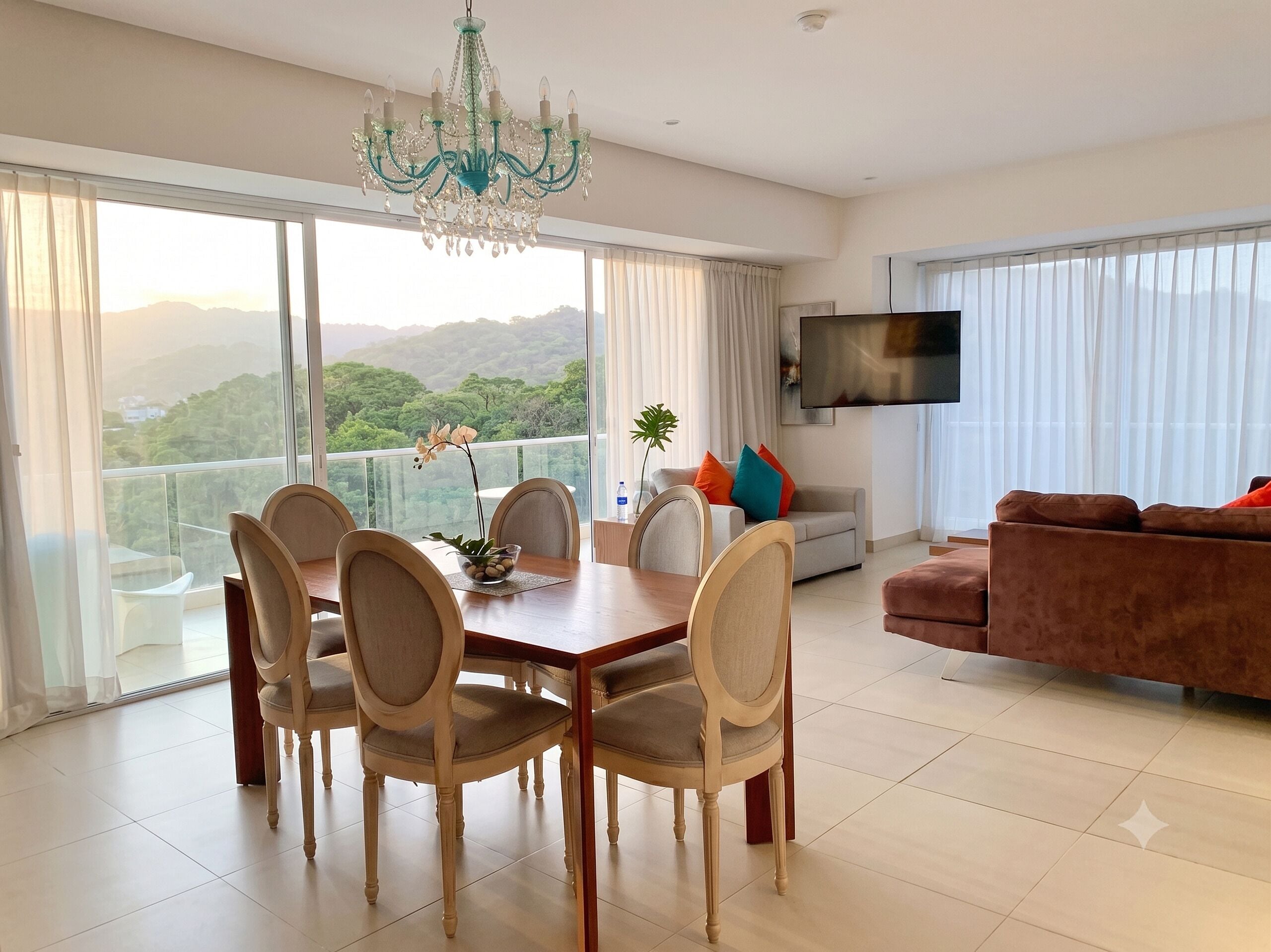 A spacious living area with a dining table, chairs, a sofa, a TV, and large windows with a view of greenery.