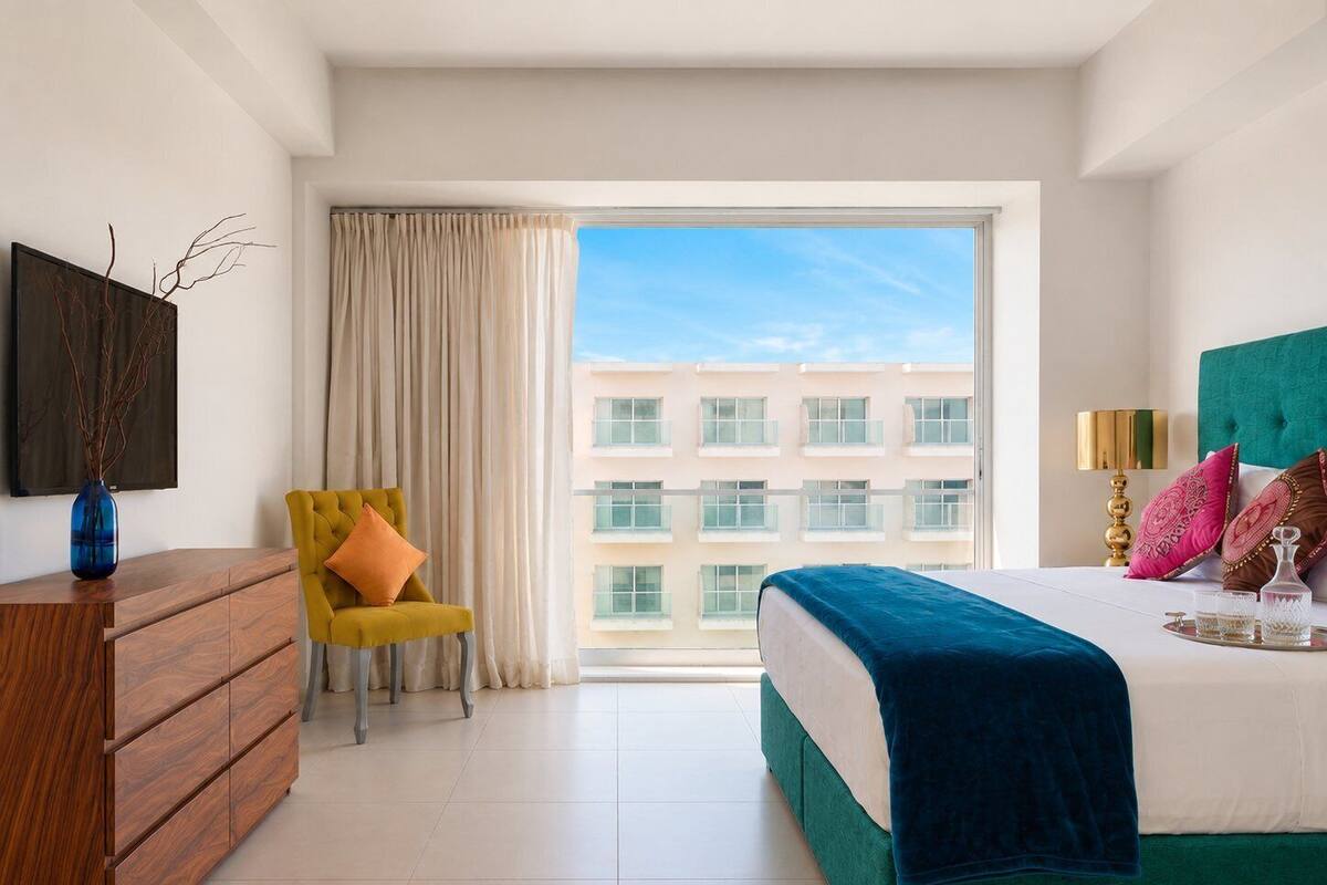 Ein modernes Hotelzimmer mit einem großen Bett, einer hölzernen Kommode, einem gelben Sessel und Blick auf ein Gebäude durch ein großes Fenster.