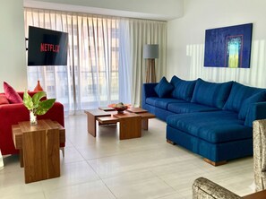 Una sala de estar moderna con un sofá azul, un sillón rojo, una mesa de centro de madera y un televisor que muestra 'NETFLIX'.