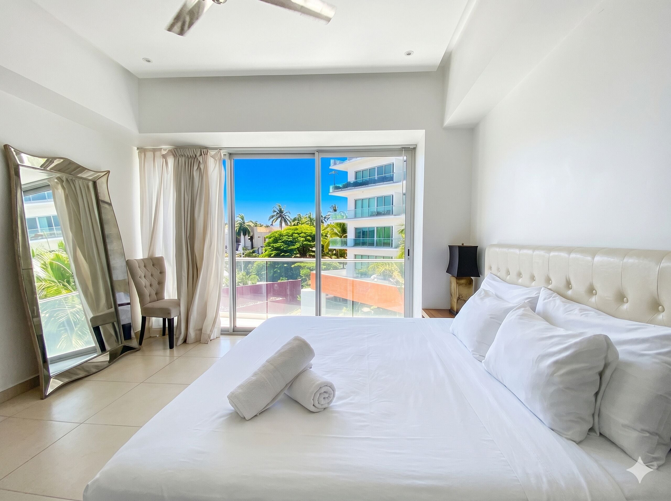A hotel room with a bed, a chair, a nightstand, a mirror, and a view of a building and greenery.