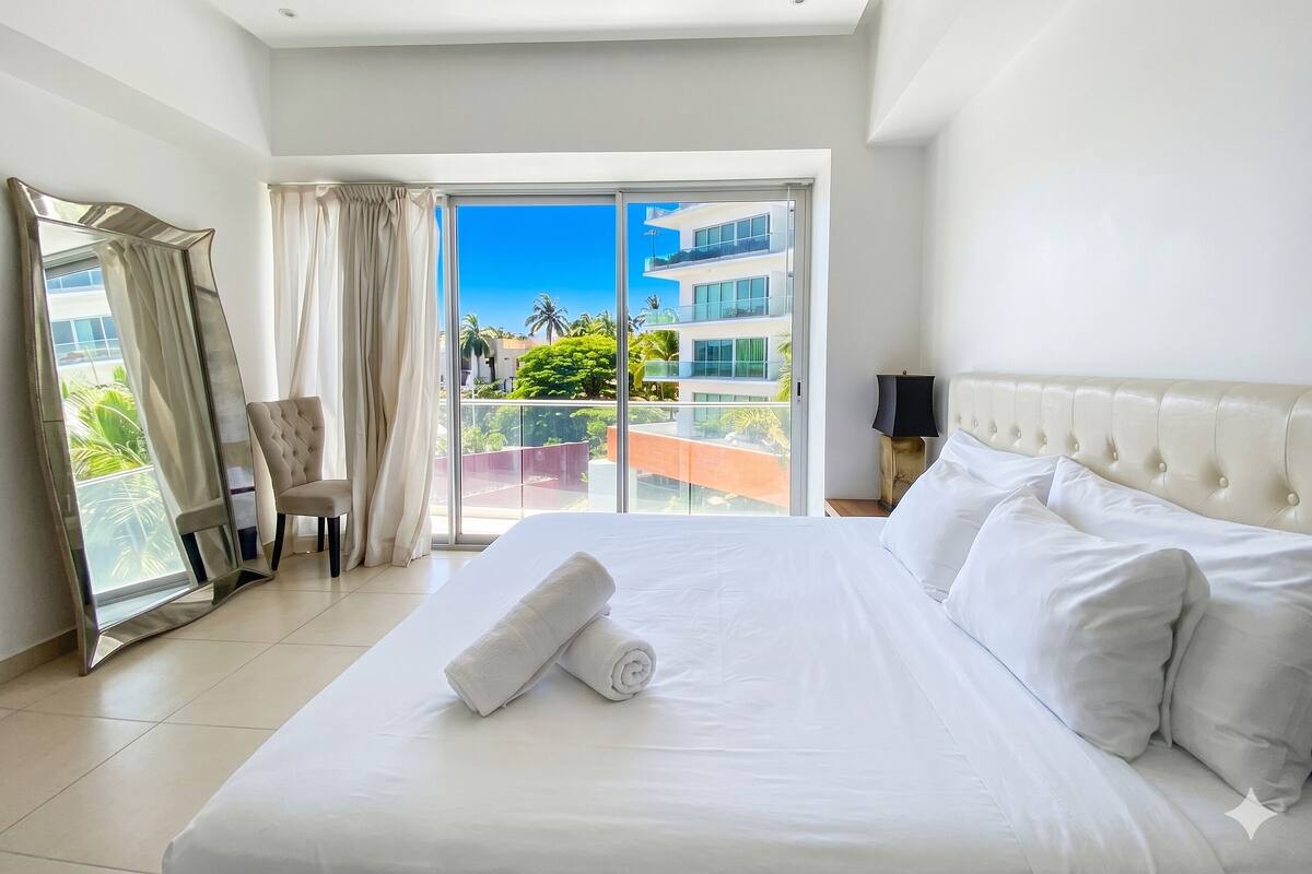 Ein Hotelzimmer mit Bett, Sessel, Nachttisch, Spiegel und Blick auf ein Gebäude und Grünflächen.