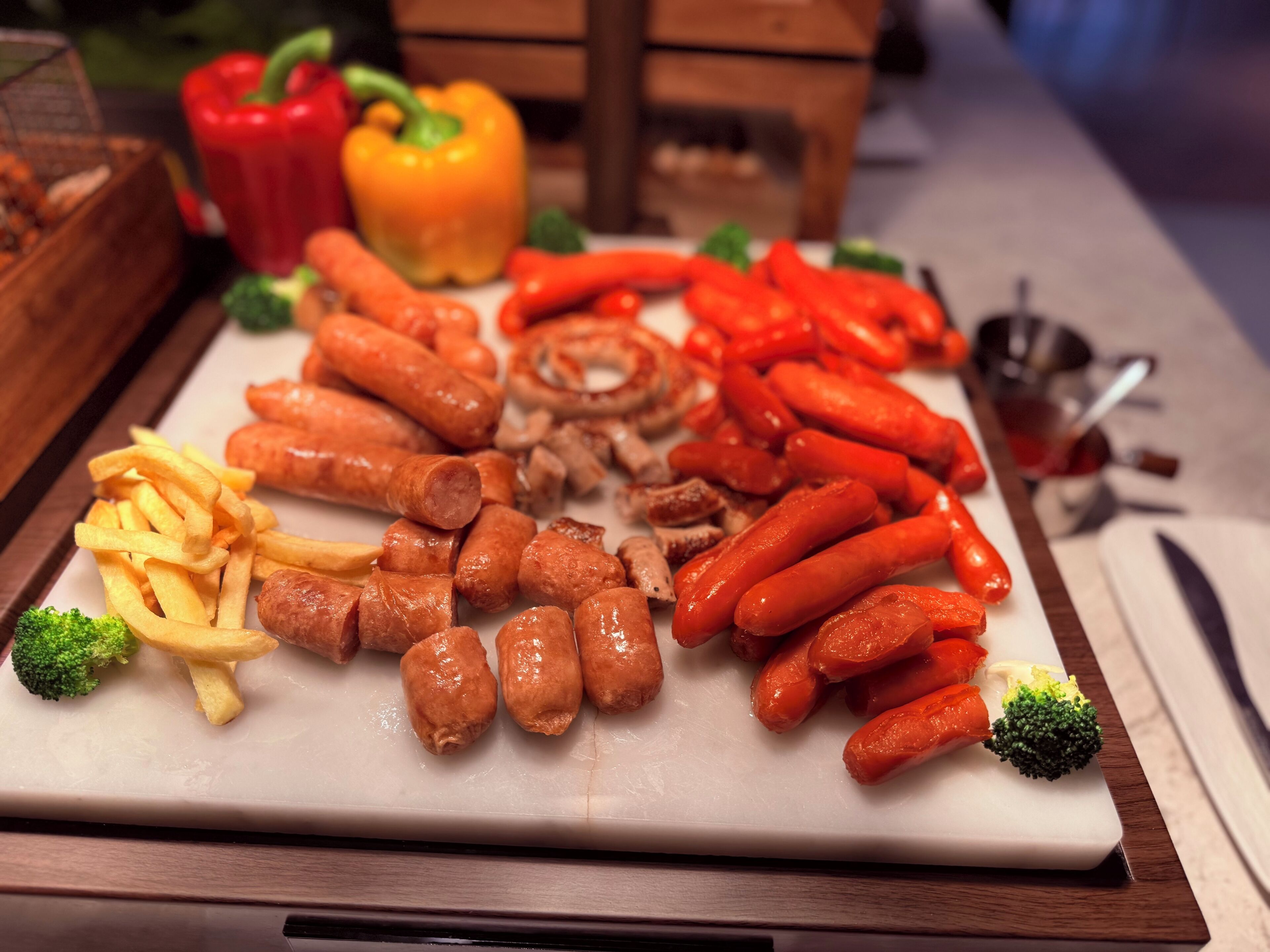 Une assiette garnie d’un assortiment de saucisses, de frites et de légumes, le tout présenté sur une planche à découper blanche.