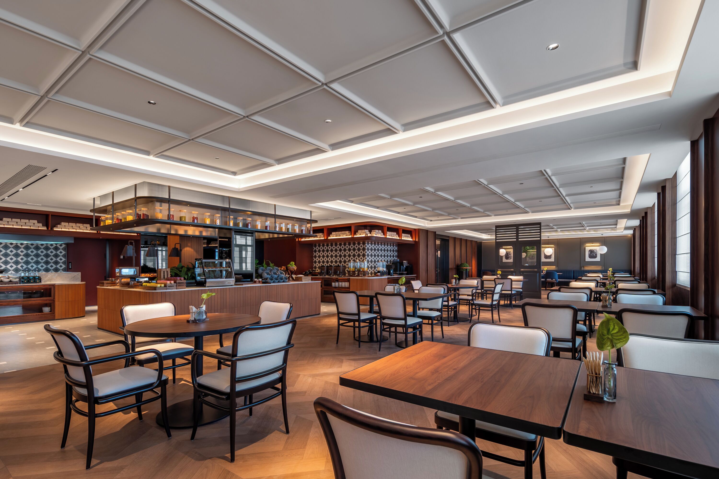 A modern restaurant with a bar area, multiple dining tables, and a high ceiling with recessed lighting.