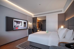 A modern hotel room with a large bed, a flat-screen TV mounted on the wall, a desk, and a walk-in closet.