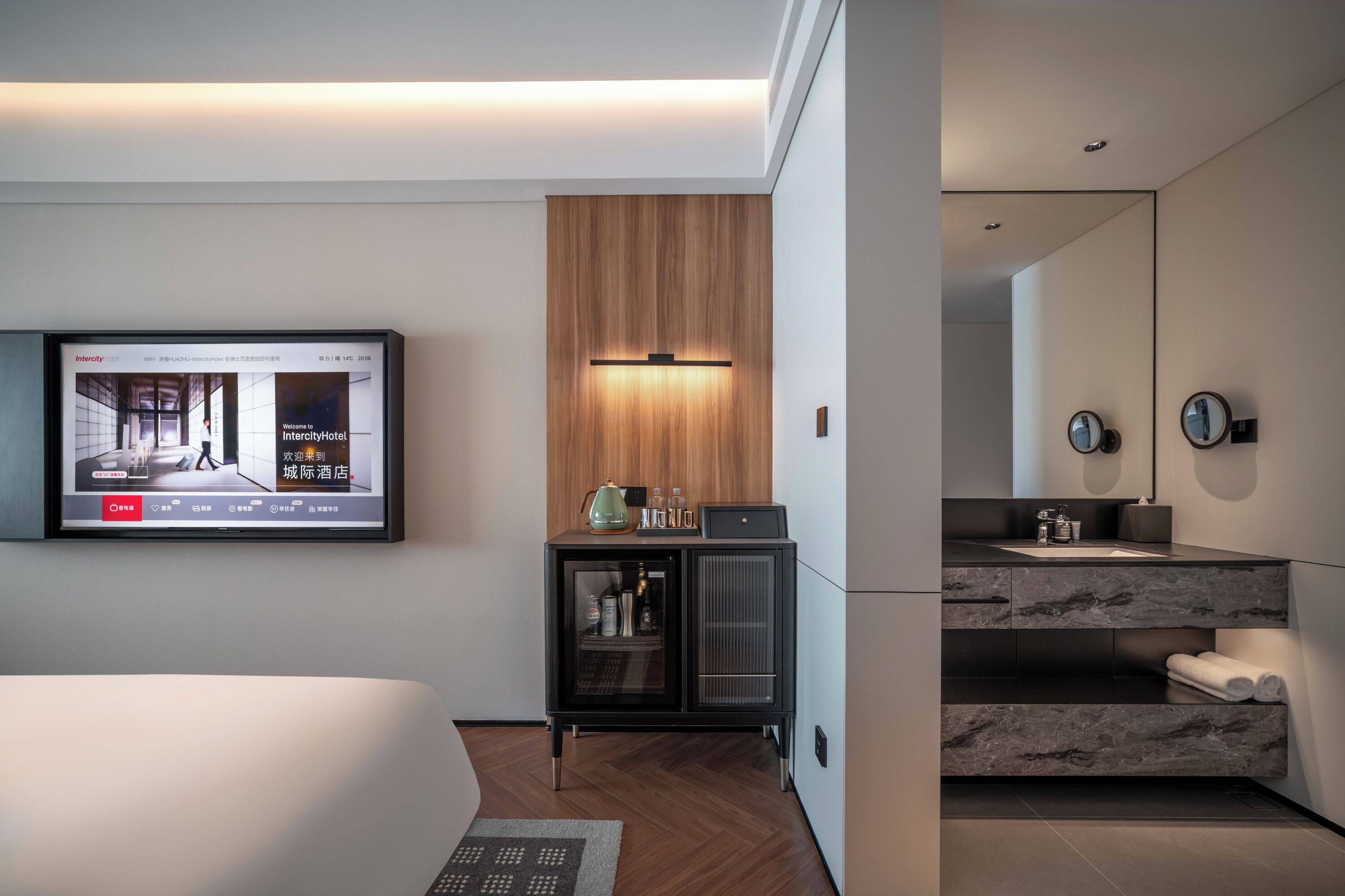 Una habitación de hotel moderna con una cama, un televisor mostrando la página web del hotel, un minibar y un baño con espejo y lavabo.