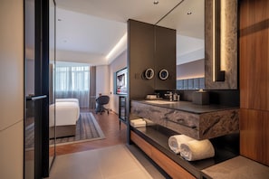Quarto de hotel moderno com um espelho grande, toucador embutido com bancada de mármore, sofá e televisão.