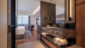 Una habitación de hotel moderna con un espejo grande, una tocador empotrado con encimera de mármol, un sofá y un televisor.