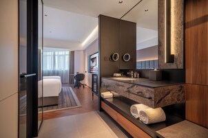 Quarto de hotel moderno com um espelho grande, toucador embutido com bancada de mármore, sofá e televisão.