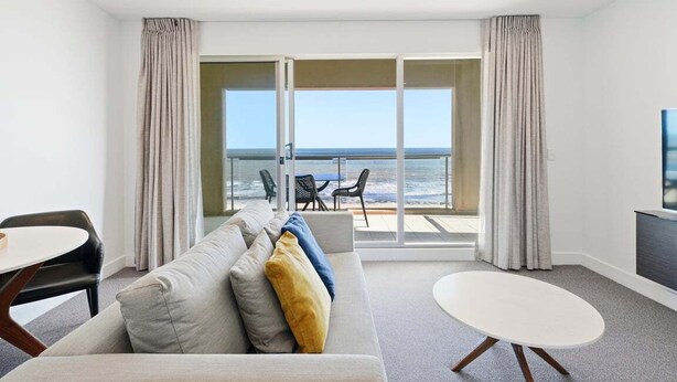 A room with a sofa, a round table, and a view of the ocean through large windows.
