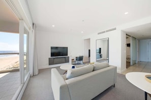 Ein modernes Wohnzimmer mit Sofa, Fernseher und Essbereich, großen Fenstern mit Blick auf den Strand und minimalistischem Design.