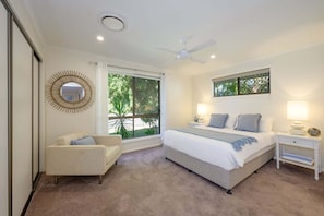 A bedroom with a bed, bedside tables, a chair, a ceiling fan, and a window with a view of greenery. - Endless Summer Stay, Noosa Heads (Noosa Heads)