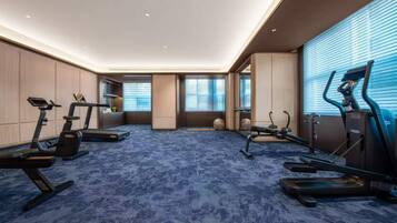 A hotel room with a blue carpet, exercise equipment, and a large window with blinds.