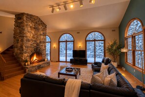 A spacious living room with a stone fireplace, a large leather sofa, a flat-screen TV, and a staircase leading to an upper level.