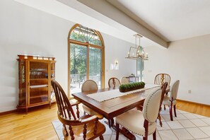 Comedor con mesa de madera, sillas, una araña y un ventanal con vistas a árboles.