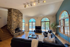 Un salón amplio con chimenea de piedra, un sofá de cuero negro y ventanas arqueadas grandes.