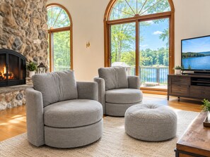 A living room with a stone fireplace, two grey armchairs, a round ottoman, a wooden coffee table, and a TV on a wooden stand.