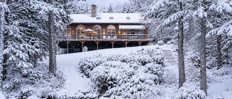 A snow-covered lodge with a deck overlooking a pond, surrounded by trees.