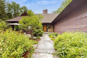 Une maison moderne avec un chemin de pierre menant à une porte en bois, le tout entouré d’une végétation luxuriante.