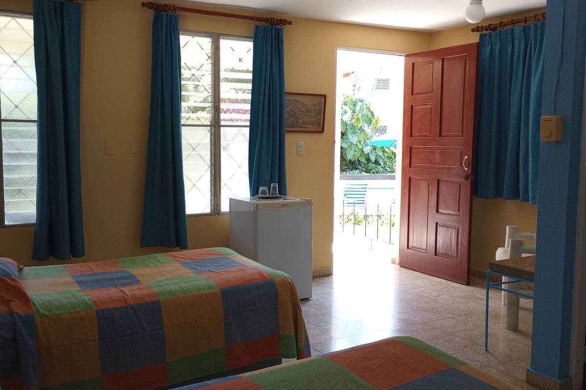 Quarto com cama, escrivaninha, frigorífico e porta para o exterior.