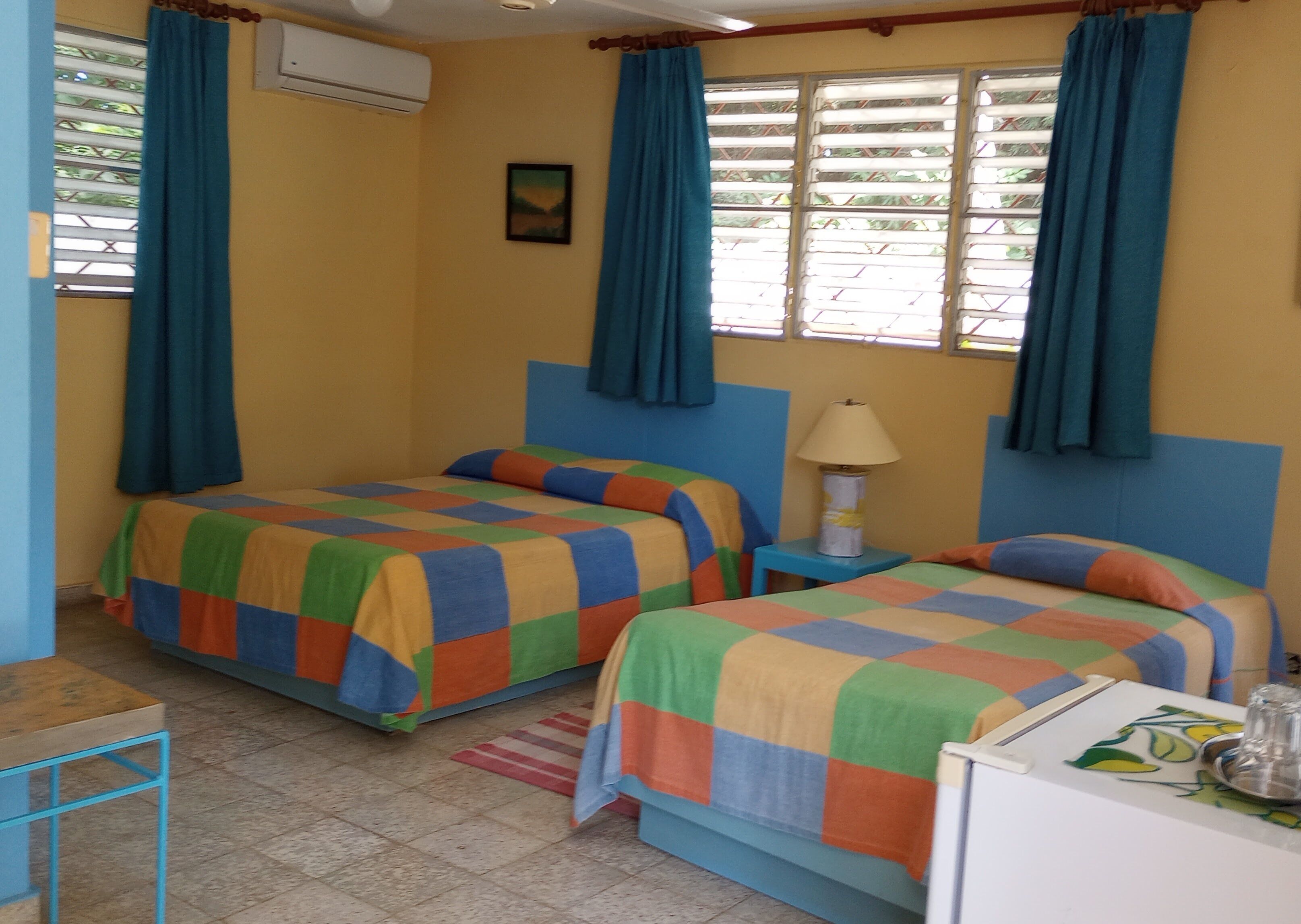 A room with two beds, a small table, a lamp, and a window with curtains.