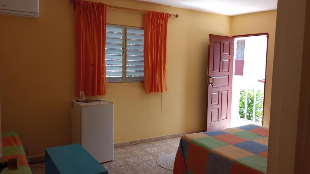 A room with a bed, a small table, a refrigerator, and a door leading to an outdoor area.