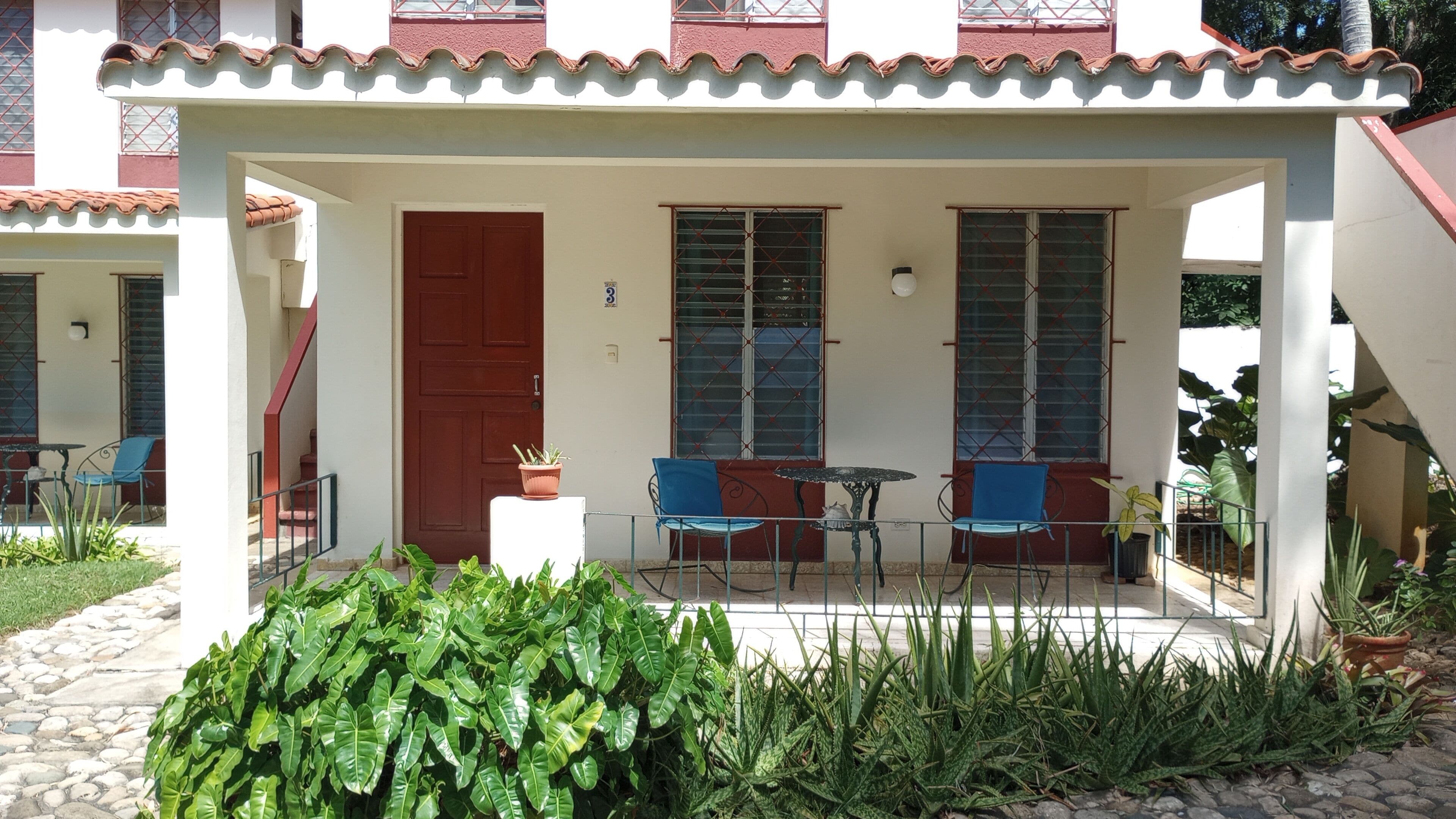 A covered outdoor area with a red door, two windows with metal bars, and blue chairs.