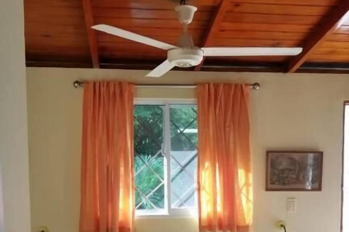 Quarto com cama, ventilador de teto, cortinas, janela e mesa de cabeceira.