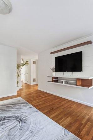 Un salon moderne avec un téléviseur à écran plat fixé à un mur blanc, un parquet en bois et un grand tapis à motifs abstraits.