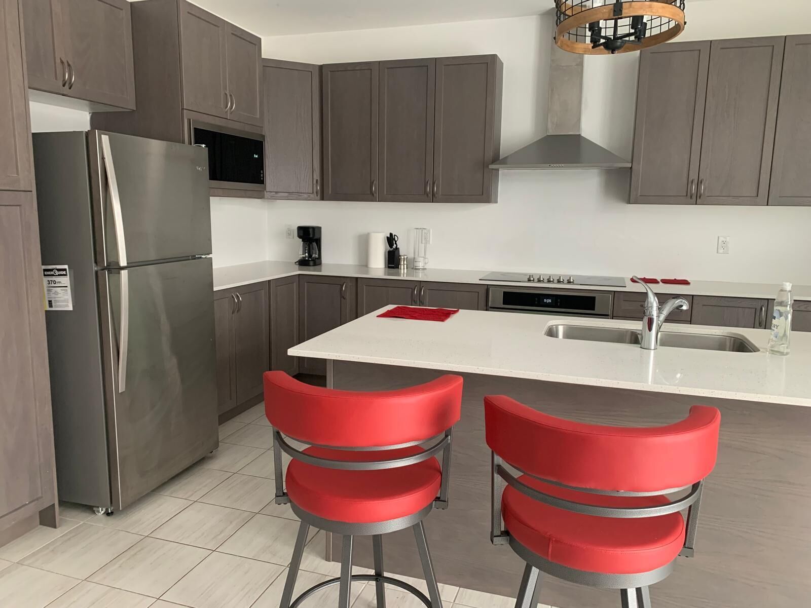 A modern kitchen with grey cabinets, a stainless steel refrigerator, a built-in microwave, a coffee maker, and a sink. There are two red bar stools at the island.. Enjoy the comfort of this cozy home, minutes away from downtown!