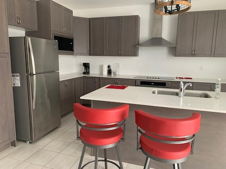 A modern kitchen with grey cabinets, a stainless steel refrigerator, a built-in microwave, a coffee maker, and a sink. There are two red bar stools at the island.. Enjoy the comfort of this cozy home, minutes away from downtown!