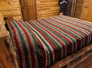 Interior d'una cabana de fusta amb un llit, parets de fusta i una persona al costat d'una porta.