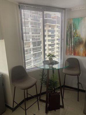 A room with a glass table, two chairs, a potted plant, a painting, and a view of buildings through blinds. - CAPSULA EXPRES PUNTA PAITILLA PANAMA/ENCHUFE WIFI WIFI TABLE TOP TYPE WORKBENCH. (Panamá)