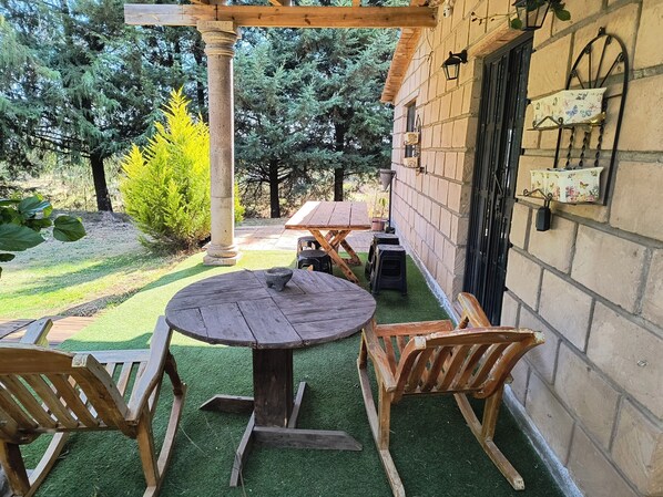 A patio with a round wooden table and chairs, a stone pillar, and a brick wall with a window and wall-mounted lights.